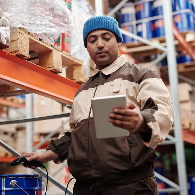 Warehouse worker in a blue beanie checking a tablet while holding a barcode scanner beside stocked shelves