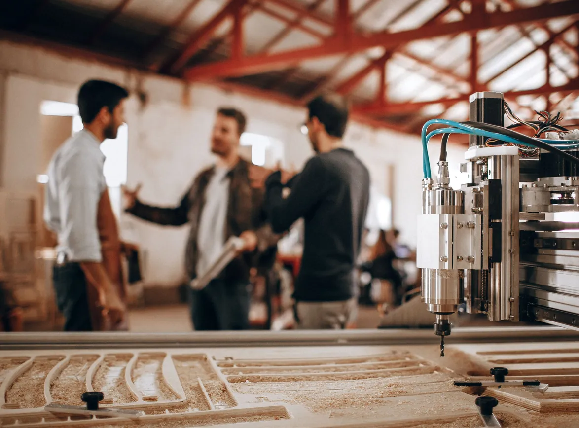 Three men talk on a workshop floor behind a CNC router cutting wooden parts