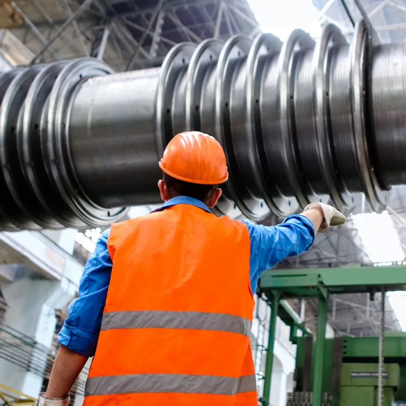 Worker in a hard hat and high-visibility vest pointing at a large turbine rotor in a factory