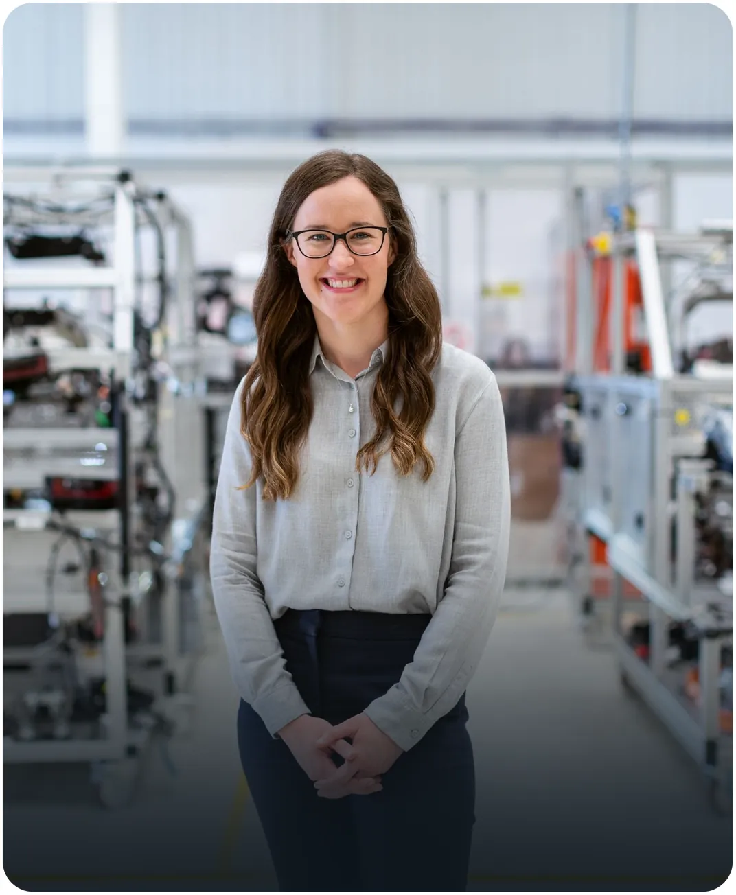 Smiling engineer in glasses standing on a factory floor with assembly line equipment behind her