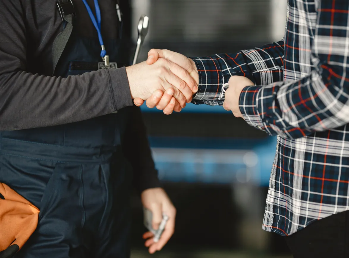 Mechanic in work overalls shakes hands with a client in a plaid shirt inside a workshop