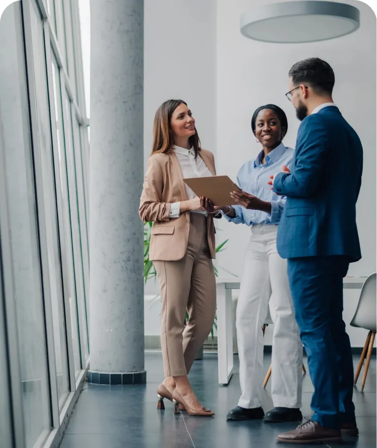 Three business colleagues talking beside tall office windows while two of them review a folder