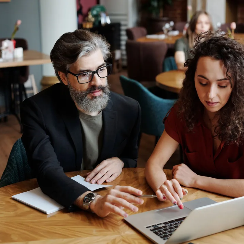 Two consultants reviewing a laptop together at a cafe table, one gesturing toward the screen