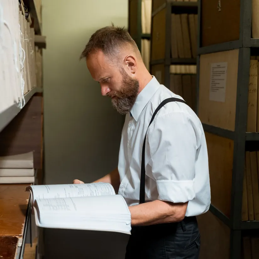 Professional reading a thick bound volume in a records archive lined with file shelves