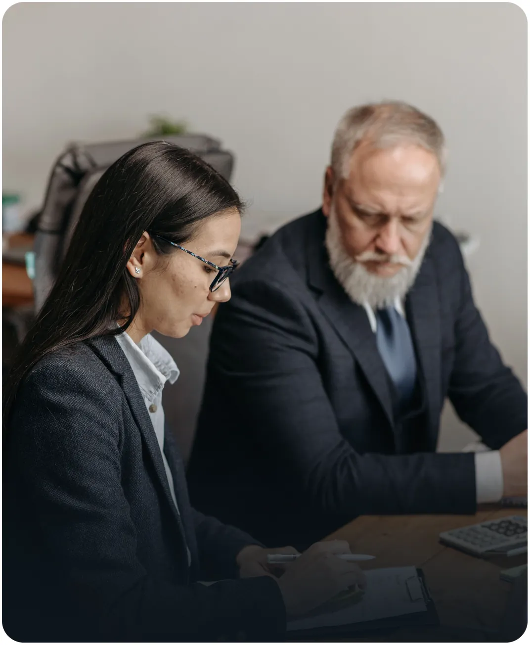 Two professionals in suits reviewing paperwork at an office desk beside a calculator