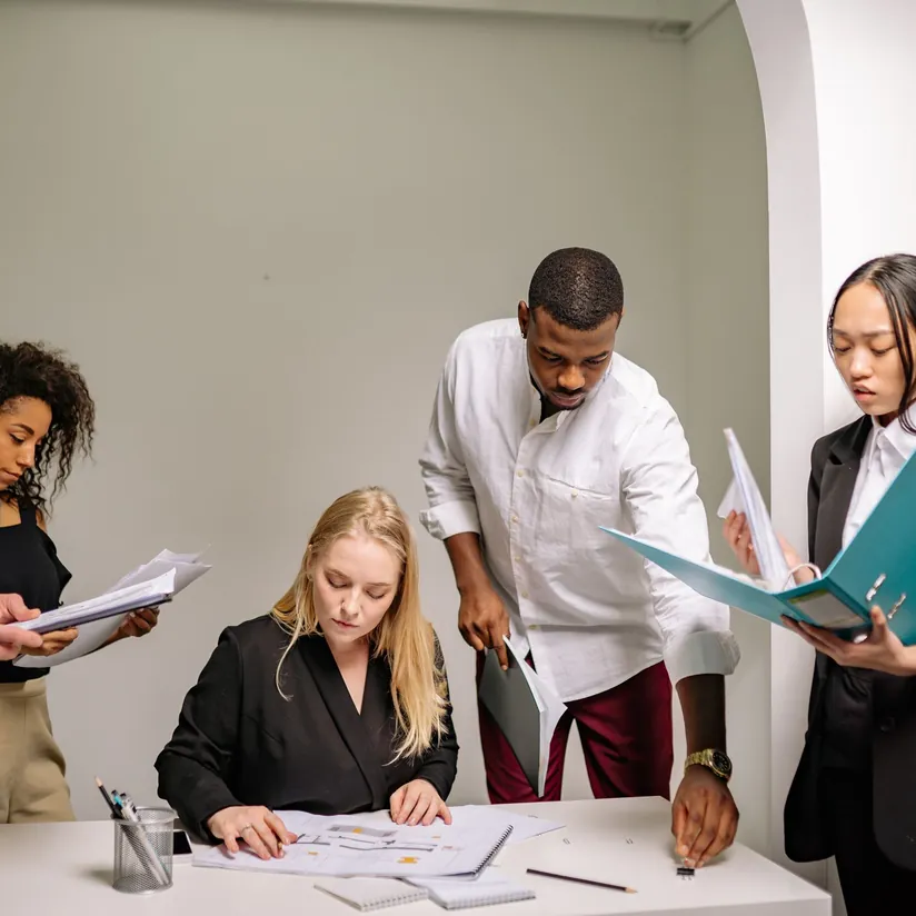 Four colleagues huddled around a desk reviewing documents, charts and binders