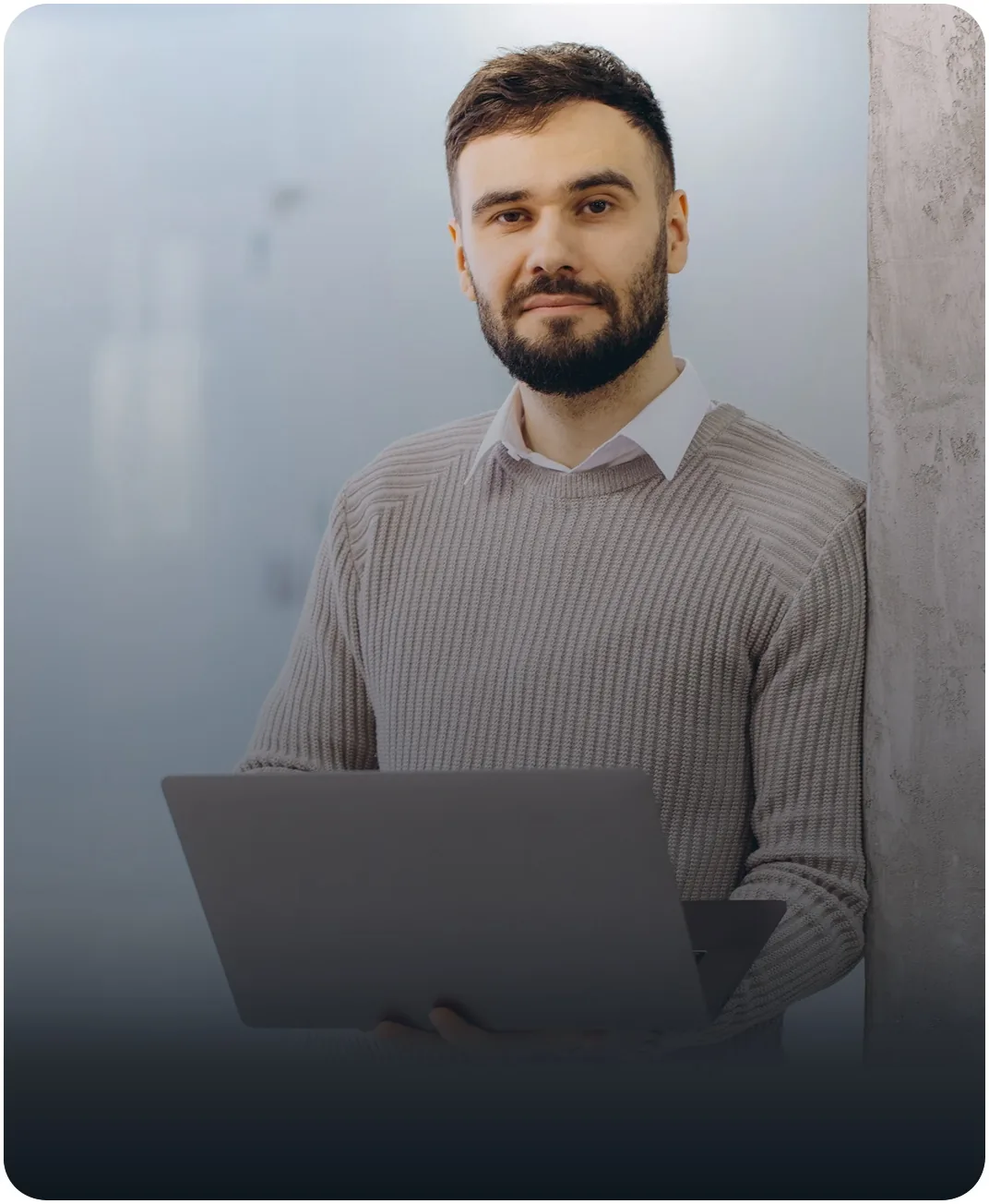 Bearded consultant in a gray sweater holding an open laptop while leaning against a concrete pillar