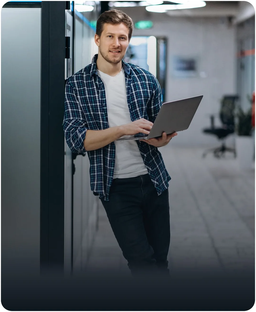 Smiling man in a plaid shirt leaning against a wall in an office hallway while typing on a laptop