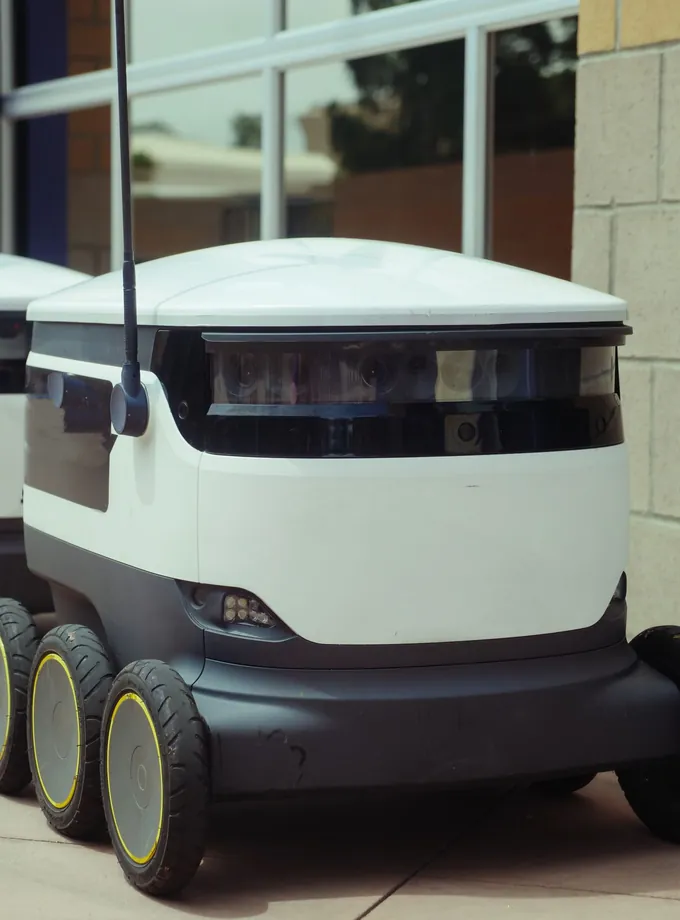 White six-wheeled autonomous delivery robot parked on a sidewalk beside a building's glass windows