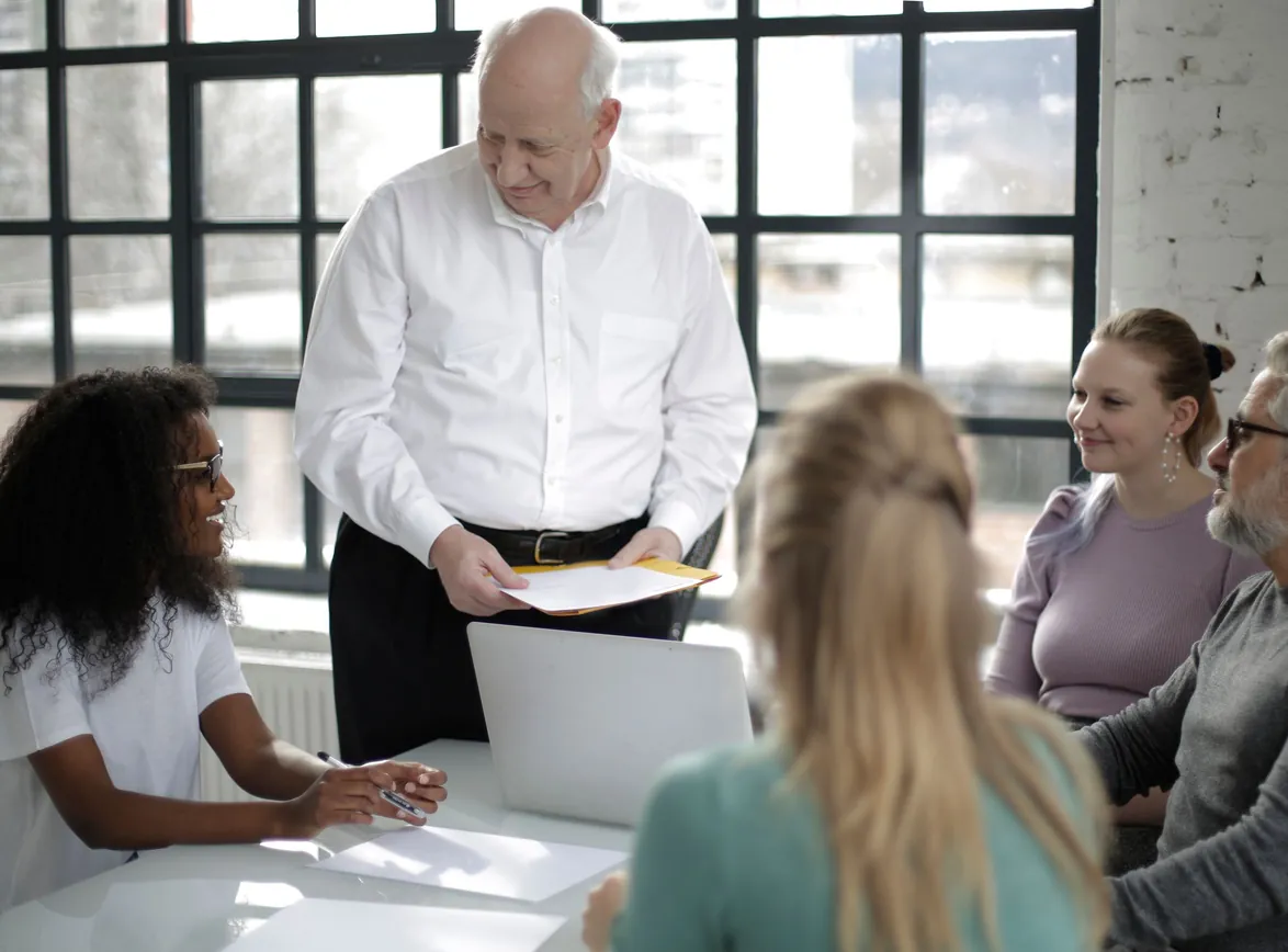 Senior advisor in a white shirt stands at a meeting table holding a folder while four team members listen